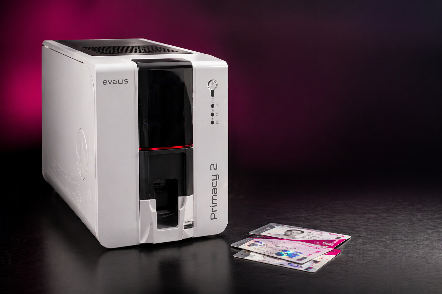 White Evolis Primacy 2 card printer with three plastic ID cards laid on a dark surface, pink spotlight in background